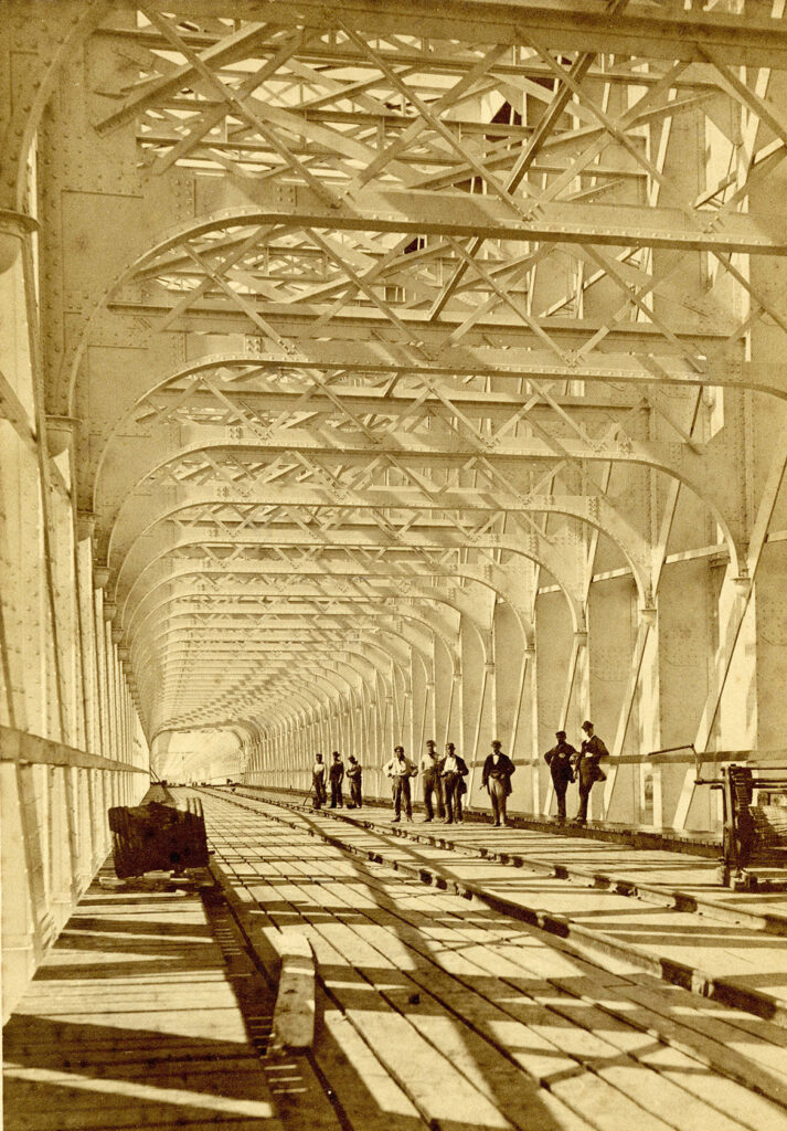 Technische hoogstandjes van de lange, kolossale spoorbrug in aanbouw over rivier de Lek in Culemborg. De afmetingen worden geaccentueerd door de kleine gestalten erin en de hoge ijzeren leggers die, dankzij het schuine standpunt, ritmisch in de verte verdwijnen.