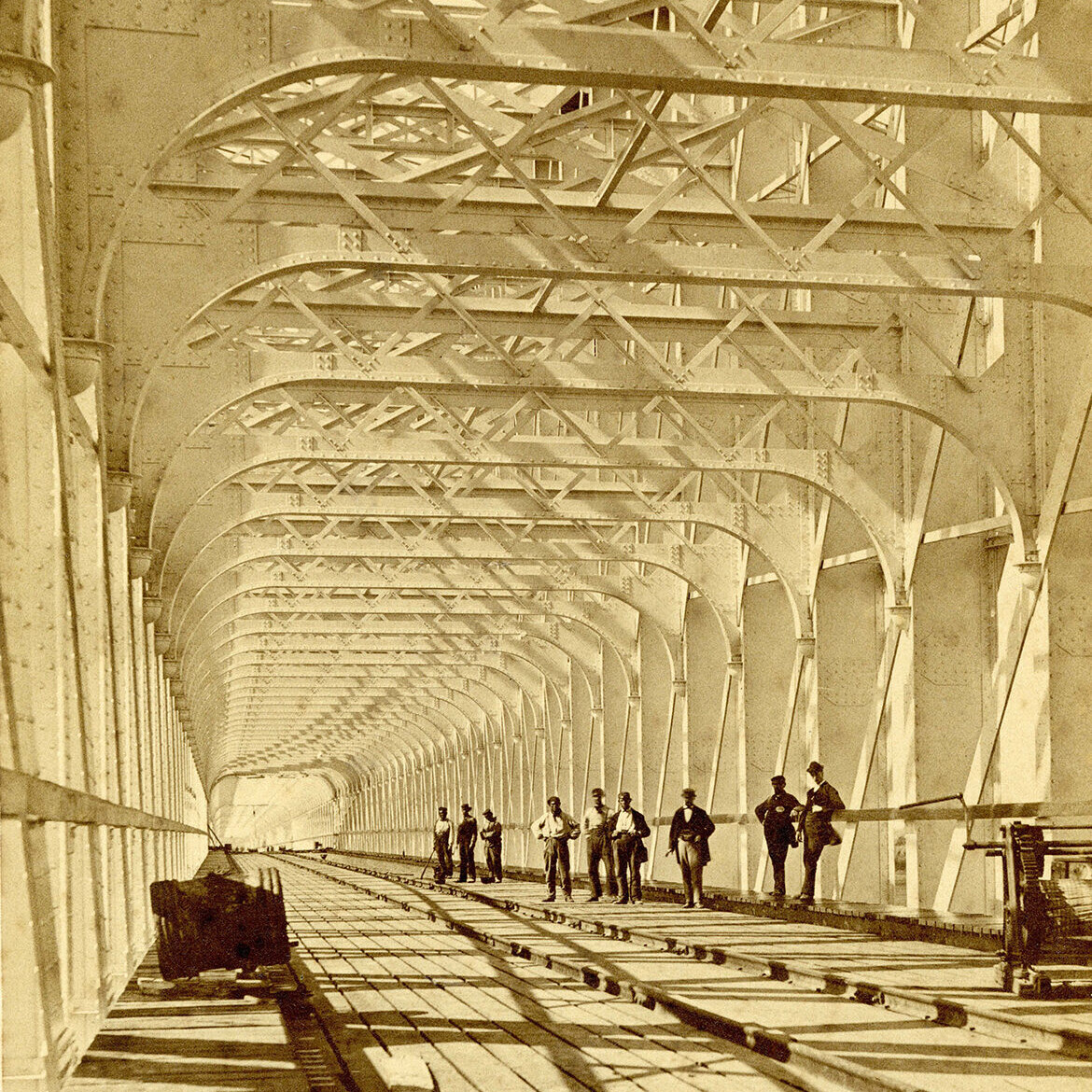 Technische hoogstandjes van de lange, kolossale spoorbrug in aanbouw over rivier de Lek in Culemborg. De afmetingen worden geaccentueerd door de kleine gestalten erin en de hoge ijzeren leggers die, dankzij het schuine standpunt, ritmisch in de verte verdwijnen.