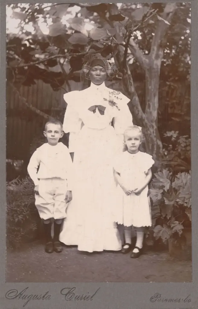 Portretfoto van een Afro-Surinaams kindermeisje met een blank jongetje en meisje voor een boom.