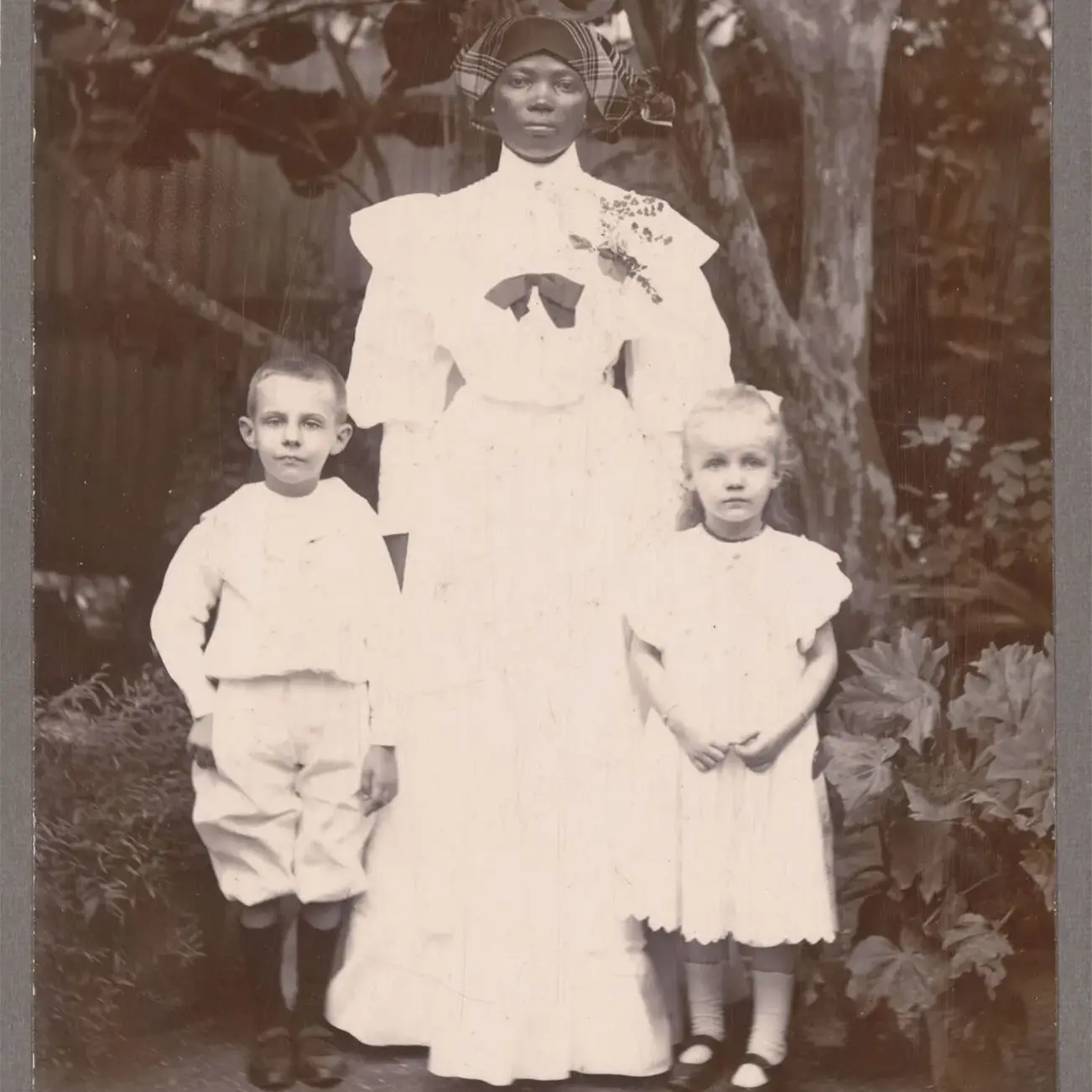 Portretfoto van een Afro-Surinaams kindermeisje met een blank jongetje en meisje voor een boom.