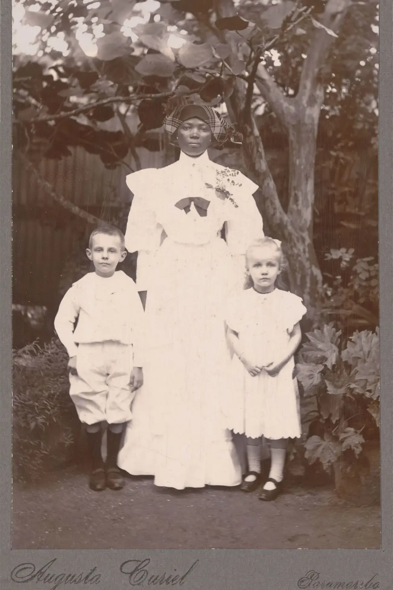 Portretfoto van een Afro-Surinaams kindermeisje met een blank jongetje en meisje voor een boom.
